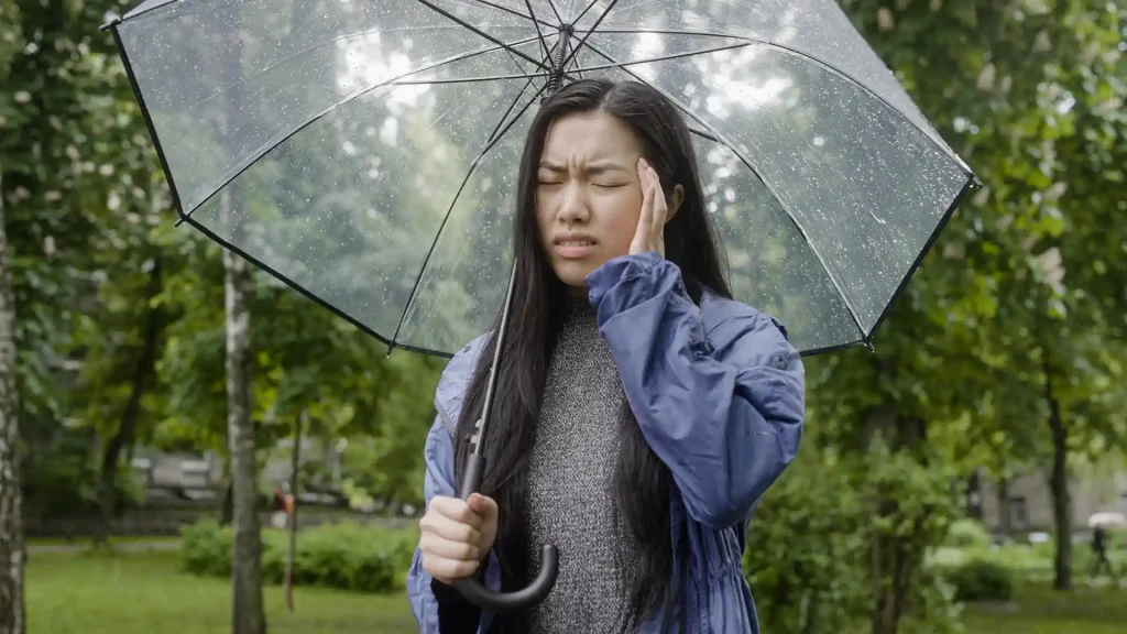 Young woman in a raincoat holding an umbrella, touching her head with a pained expression in a park