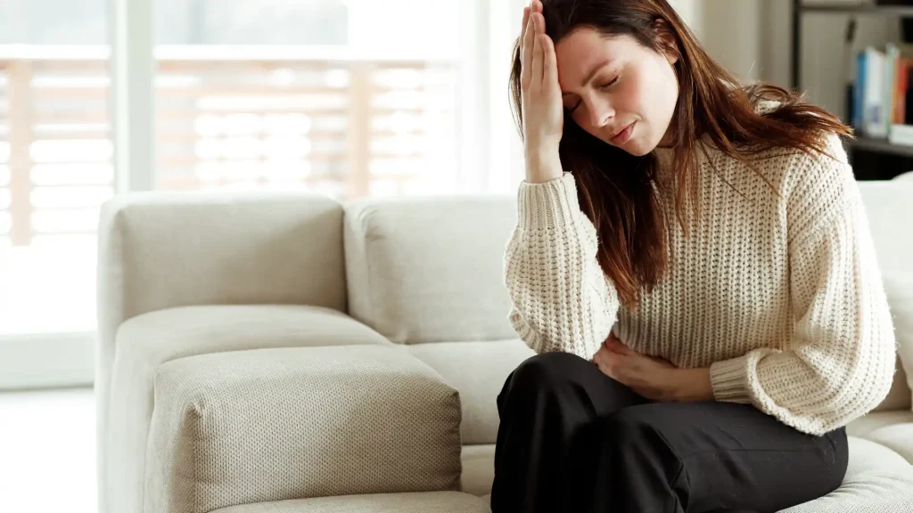 A woman sitting on a couch, holding her head with one hand, looking distressed or overwhelmed. She appears to be experiencing discomfort or pain.